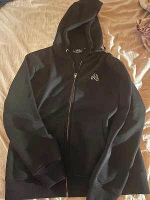 Svart hoodie med dragkedja från Maggiore - Snygg svart hoodie från Maggiore med dragkedja framtill och justerbar huva. Diskret logga broderad på bröstet och ribbade muddar vid ärmar och nederkant. Perfekt för en chill och stilren look. Materialet känns mjukt och bekvämt.