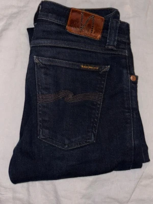 Mörkblå jeans från Nudie Jeans - Säljer ett par mörkblå jeans från Nudie Jeans i slim, med klassisk femficksmodell och snygga kontrastsömmar på bakfickan. Jeansen har en skinnpatch med logga i midjan och är tillverkade i slitstarkt denimtyg. Perfekta för dig som gillar stilrena och tidlösa jeans.  Jeansen är i storlek 27/30