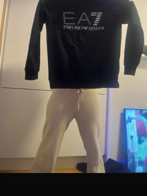 Svart EA7 Emporio Armani hoodie - Säljer en svart hoodie från EA7 Emporio Armani med stor logga framtill i vitt och silver. Klassisk modell med långa ärmar och rund halsringning. Perfekt för dig som gillar stilrena och exklusiva plagg.den är till både kvinnor och män. 