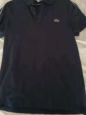 Mörkblå pikétröja från Lacoste - Klassisk mörkblå pikétröja från Lacoste med den ikoniska gröna krokodilloggan på bröstet. Tröjan har krage, knappar vid halsen och vita ränder på axlarna. Regular fit och tillverkad i mjuk bomull som känns skön mot huden.