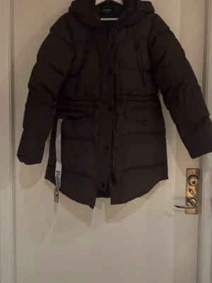 Mörkbrun vinterjacka med huva - Vinterjacka från Everest med huva (Modell: W Warm Parka) i färgen Plum Slate. Fyra fickor framtill och även en innerficka. Kan dras åt i midjan. Jackan är i nyskick och använd fåtal gånger, säljes pågrund av för liten storlek. Storlek 36 (liten i storlek). Nypris 1499kr