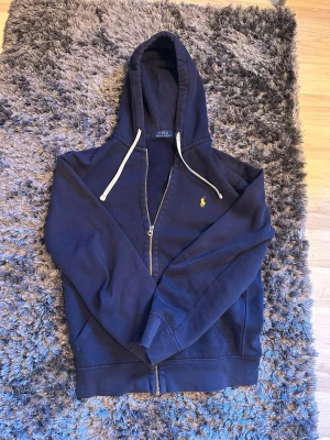 Mörkblå hoodie från Polo Ralph Lauren - Mörkblå hoodie med dragkedja från Polo Ralph Lauren. Klassisk design med vit dragsko i huvan och gul broderad logga på bröstet. Tillverkad i mjukt bomullsmaterial och har kängurufickor framtill. Perfekt för en avslappnad och stilren look.
