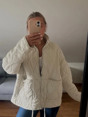 Beige quiltad vindjacka med dragkedja - Säljer en beige quiltad vindjacka med dragkedja framtill och två stora fickor. Jackan har en avslappnad passform, hög krage och dragsko i nederkant. Materialet är mjukt och lätt, perfekt för lager på lager under kyliga dagar.