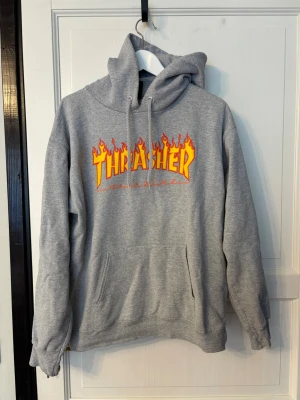 Thrasher hoodie (S) - Säljer en grå hoodie från Thrasher med den ikoniska flames-loggan i gult och rött på bröstet. Hoodien har huva med dragsko och en stor magficka. Perfekt för dig som gillar streetwear och skatekultur.