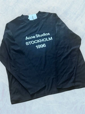 Svart långärmad topp Acne Studios - Svart långärmad topp från Acne Studios med vit text 'Acne Studios STOCKHOLM 1996' på bröstet. Klassisk rak passform och rund halsringning. Perfekt till jeans eller kjol för en clean och stilren look.