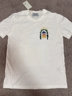 Vit Casablanca t-shirt med tryck - Snygg vit t-shirt från Casablanca med färgglatt tryck av en byggnad och texten 'Avenida Casablanca' på bröstet och ryggen. T-shirten har korta ärmar och är gjord i mjuk bomull. Perfekt för dig som gillar stilrena plagg med unika detaljer.