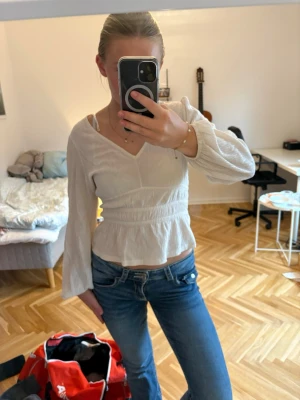 Vit blus med v-ringning och resår - Säljer en söt vit blus med v-ringning och långa ärmar. Blusen har resår i midjan som ger en snygg passform och lätt volang nertill. Perfekt till jeans eller kjol för en fräsch look.