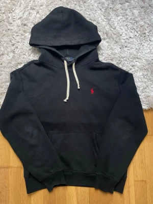 Svart hoodie från Polo Ralph Lauren - Säljer en svart hoodie från Polo Ralph Lauren med klassisk röd logga broderad på bröstet. Hoodien har vita dragsnören, stor huva och en magficka. Tillverkad i mjukt bomullsmaterial, perfekt för en chill och stilren look. Lite sliten vid ena mudden 