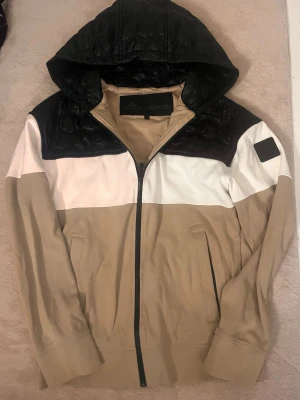 Moose Knuckles hoodie jacka - Snygg vindjacka från Moose Knuckles i beige, vitt och svart. Jackan har quiltad huva och axelparti, dragkedja framtill och två fickor. Patch med logga på ärmen ger extra stilpoäng. Perfekt för dig som vill ha en streetinspirerad look.