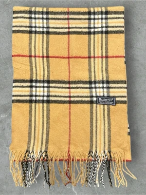 Beige rutig halsduk från Burberry - Klassisk beige halsduk från Burberry med ikoniskt rutmönster i svart, vitt och rött. Halsduken har fransar i ändarna och är tillverkad i mjukt material som känns lyxigt och stilrent. Perfekt accessoar för att lyfta din outfit.