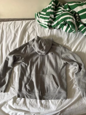 Grå Polo Raulph Lauren kofta  - Säljer en stilren Polo Raulph Lauren grå zipup hoodie till original pris vid 1000kr. Hoodien har två stora fickor framtill och mjukt, luddigt innerfoder. Perfekt för chill dagar eller när du vill ha något bekvämt. Enkel och klassisk design som passar till det mesta. Lite oversized vid ärmarna. 
