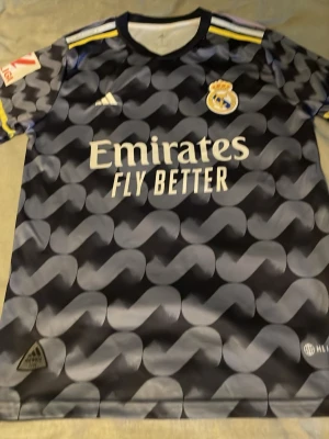 Real Madrid bortatröja Vini Jr 7 - Snygg Real Madrid bortatröja från Adidas med Vini Jr och nummer 7 på ryggen. Tröjan är svart med grått mönster, gula detaljer på axlarna och tryck. Tillverkad i lätt och ventilerande material, perfekt för fotboll eller träning.