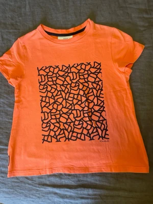 Orange t-shirt från Johaug NOW stl.XS - Stl.XS, Snygg orange t-shirt från Johaug NOW i bomull med ett svart abstrakt tryck på framsidan. Släppt sömn nere pp ryggen runt 1 cm (syns på bild 3-4)