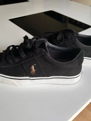 Svarta Polo Ralph Lauren sneakers - Svarta låga sneakers från Polo Ralph Lauren med klassisk broderad logga på sidan och POLO-text på hälen. Skorna har svart canvasmaterial, svarta skosnören och vit sula med svart rand. Perfekta för en clean och stilren look. Liten 38 så skulle säga strl 37.