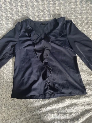 Mörkblå blus med volangkant - Stilren mörkblå blus med långa ärmar och snygg volangkant längs den djupa v-ringningen. Blusen har en lätt glansig yta och är figurnära i modellen. Perfekt för dig som gillar en chic och enkel look.