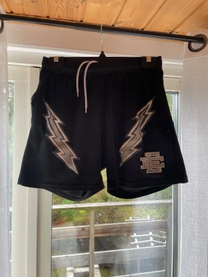 Svarta shorts med blixtar och snörning - Säljer ett par svarta shorts med meshstruktur och vita snören i midjan. Shortsen har två stora blixtmotiv i silver på framsidan och en broderad logga i matchande färg. Perfekta för dig som vill sticka ut med en cool och sportig stil.