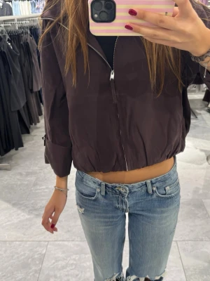 Mörkbrun bomberjacka med dragkedja - Snygg mörkbrun bomberjacka med dragkedja framtill och resår i midjan. Jackan har en avslappnad passform och långa ärmar. Perfekt för dig som vill ha en stilren och trendig look.
