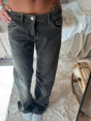 Baggy grå jeans från Zara - Säljer ett par baggy grå jeans från Zara i storlek 36. Jeansen är lågmidjade. Jeansen har en avslappnad passform, fem fickor och klassisk knappgylf. Materialet är denim med slitna detaljer och ljusa tvättade partier för en cool vintage-look.