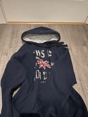 Mörkblå hoodie med London-tryck - Mörkblå hoodie med stor huva och vitt innerfoder. Framsidan har ett slitet tryck med texten 'THIS IS LONDON' och en Union Jack-flagga i rött och vitt. Hoodien har känguruficka och dragsko i huvan. Perfekt för dig som gillar streetwear och brittisk stil.