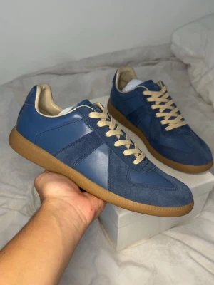 Maison Margiela - blå - Snygga blå sneakers från Maison Margiela med beige detaljer och gummisula. Skorna har en mix av mocka och läder, beige snörning och klassisk låg siluett. Perfekta för dig som gillar stilrena och trendiga sneakers.
