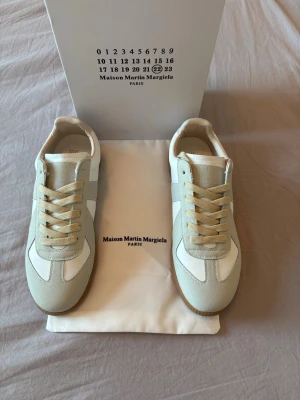 Maison Margiela vita sneakers - Stilrena sneakers från Maison Martin Margiela i vitt och beige med klassisk gummisula. Ovandelen är i en mix av mocka och slätt läder, med beige snörning och diskreta detaljer. Perfekta för dig som gillar minimalistisk och trendig design.