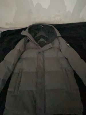 Grå dunjacka med huva - Säljer en grå dunjacka med huva och två stora fickor framtill. Jackan har en klassisk puffer-design och en patch på ena ärmen. Perfekt för kalla vinterdagar när du vill hålla dig varm och stilsäker.