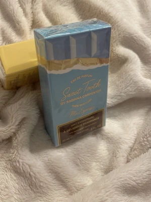 Sweet Tooth Eau de Parfum 30 ml - Sweet Tooth Eau de Parfum från Sabrina Carpenter i en snygg blå och guldig förpackning. Doften är på 30 ml och beskrivs som en läcker och söt parfym. Förpackningen har en modern, rektangulär form och är inslagen i plast.