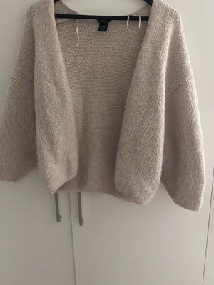 Beige stickad kofta med lös passform - Mysig beige stickad kofta med öppen front och avslappnad, boxig passform. Koftan har långa ärmar och är tillverkad i ett mjukt, grovt garn som ger en skön känsla. Perfekt att slänga över en t-shirt eller topp för extra värme.
