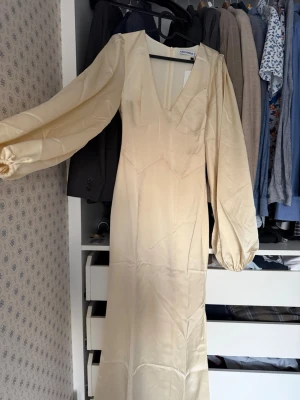 Adoore klänning  - Elegant långklänning i champagnefärgad satin med djup v-ringning och långa puffärmar med resår vid handlederna. Klänningen har en smickrande passform och ett stilrent, minimalistiskt uttryck. Perfekt för dig som vill sticka ut med en lyxig look.