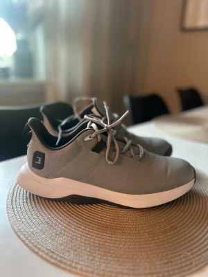 Footjoy Golfskor - Snygga grå sneakers med vit sula och svarta detaljer. Skorna har en låg profil och snörning framtill. Materialet ser ut att vara en mix av syntet och textil, vilket ger en modern och sportig look. Perfekta för dig som gillar stilrena och bekväma skor. Perfekt för golf, Använda 2 rundor