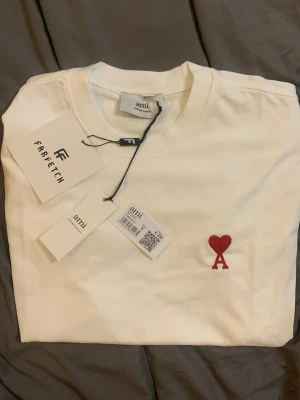 Vit t-shirt från Ami med rött hjärta - Säljer en vit t-shirt från Ami med ett broderat rött hjärta och bokstaven A på bröstet. Klassisk rund halsringning och korta ärmar. Tillverkad i mjuk bomull, perfekt för en clean och stilren look.