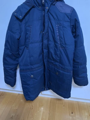 Blå dunjacka med huva - Säljer en blå dunjacka med huva och flera fickor framtill. Jackan har quiltad design, dragkedja och tryckknappar samt mörkare detaljer på axlar och ärmar. Perfekt för kalla vinterdagar och har en avslappnad passform.