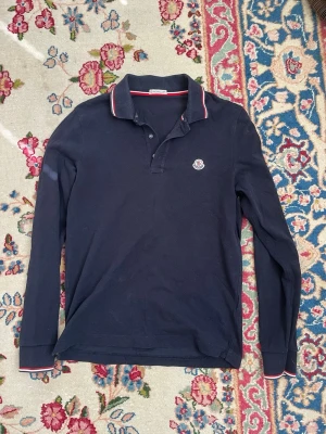 Mörkblå långärmad pikétröja Moncler - Snygg mörkblå långärmad pikétröja från Moncler med klassisk krage och knappar framtill. Tröjan har Moncler-logga på bröstet och diskreta röda och vita detaljer på krage och ärmslut. Perfekt för dig som gillar stilrena plagg.
