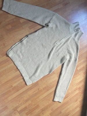 Beige stickad tröja med halv dragkedja - Säljer en beige stickad tröja med turtle neck och halv dragkedja med ringdetalj. Tröjan har ett grovt stickat mönster, långa ärmar och slits på båda sidor längst ner. Den är i bra skick och enkel att styla till många olika saker!!