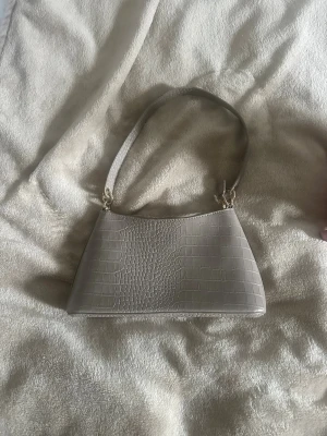 Beige axelremsväska med krokoprägling - Stilren beige handväska med axelrem och krokodilpräglat mönster. Väskan har en avlång, lätt utsvängd form och stängs med dragkedja. Perfekt accessoar för att lyfta din outfit med en trendig touch.