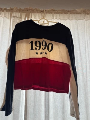 Randig långärmad topp 1990 - Cool långärmad topp med trycket '1990' och tre stjärnor framtill. Toppen har breda ränder i svart, vitt och rött och är croppad i modellen. Perfekt för dig som gillar retrostil och vill sticka ut.