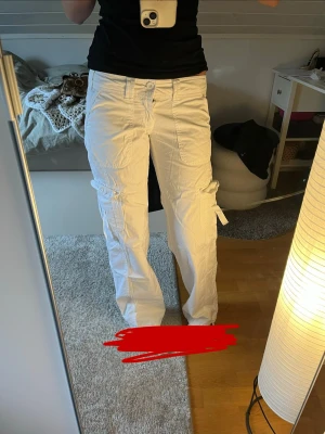 Vita cargobyxor med fickor - Säljer ett par vita cargobyxor med flera stora fickor på sidorna och bak. Byxorna har raka ben och stängs med knapp och dragkedja. Tillverkade i ett lätt tyg som känns bekvämt och passar till många olika stilar.