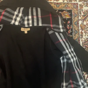 Svart hoodiejacka från Burberry - Svart hoodiejacka från Burberry med dragkedja och klassiskt rutigt foder i huvan. Jackan har en diskret Burberry-logga på bröstet och är tillverkad i mjukt material. Perfekt för dig som gillar stilrena och exklusiva plagg.