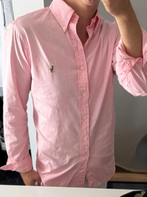 Rosa skjorta med knappar - Snygg rosa skjorta med klassisk krage och knäppning framtill. Skjortan har långa ärmar och en broderad logga på bröstet. Perfekt för en stilren look och passar till många olika outfits. Lite urtvöttad annars bra skick. 