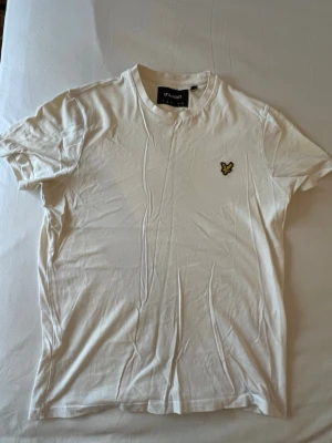 Vit t-shirt från Lyle & Scott - Klassisk vit t-shirt från Lyle & Scott med gul logga på bröstet. T-shirten har rund halsringning och korta ärmar. Tillverkad i mjuk bomull för en skön känsla och enkel stil som funkar till allt.