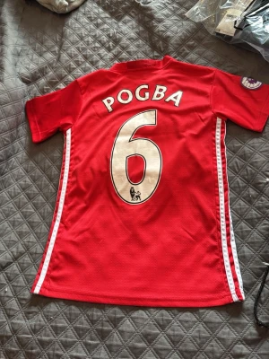 Manchester United Pogba matchtröja - Röd Manchester United matchtröja med Pogba och nummer 6 på ryggen. Tröjan har vita detaljer längs sidorna, klubbmärke och Adidas-logga på bröstet samt Chevrolet som sponsor. Tillverkad i funktionsmaterial (climacool) och har korta ärmar.