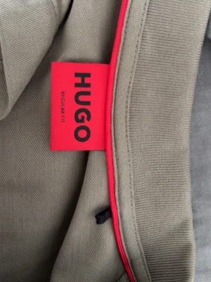 Beige pikétröja från HUGO boss - Snygg beige pikétröja från HUGO med klassisk krage och korta ärmar. Regular fit-modell i mjukt bomullsmaterial. Röd detalj på insidan av kragen ger en extra touch. Perfekt till både jeans och chinos.