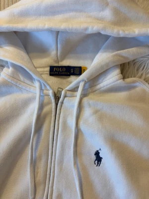 Vit hoodie från Polo Ralph Lauren - Klassisk vit hoodie från Polo Ralph Lauren med dragkedja framtill, huva med dragsko och känguruficka. Ikoniska blå loggan broderad på bröstet. Tillverkad i mjukt bomullsmaterial som är skönt att ha på sig.