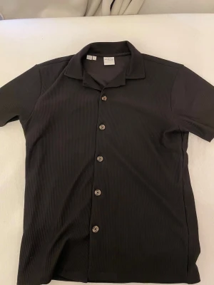 Svart ribbad skjorta från Selected Homme - Säljer en svart kortärmad skjorta från Selected Homme. Skjortan har ribbad struktur, klassisk krage och knappar framtill. Perfekt för dig som gillar stilrena och enkla plagg med en twist. Passar till många olika outfits.