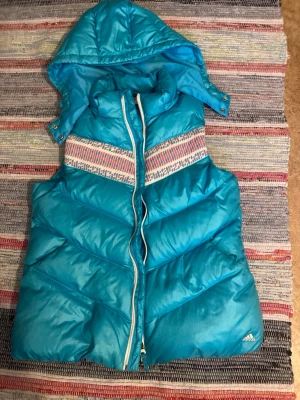 Turkos dunväst från Adidas med huva - Turkos dunväst från Adidas med avtagbar huva och dragkedja framtill. Västen har ett färgglatt mönster över bröstet och diskret Adidas-logga nedtill. Snyggt vadderad och perfekt för lager-på-lager-stil. Materialet är glansigt och ger en sportig vibe. Finns en mindre fläk på framsida. 