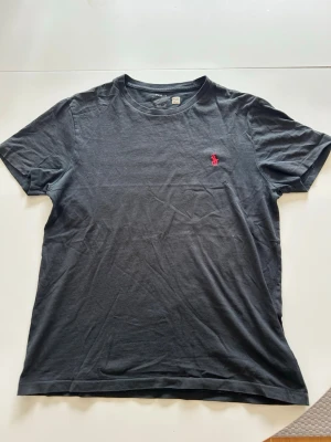 Svart t-shirt med röd logga - Snygg svart t-shirt i bomull med klassisk rund hals och korta ärmar. På bröstet finns en broderad röd logga som ger en stilren touch. Perfekt basplagg för en enkel och clean look.