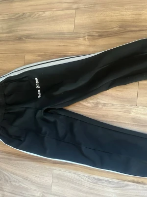 Svarta Palm Angels mjukisbyxor - Säljer ett par svarta mjukisbyxor från Palm Angels med vita sidoränder och broderad logga på vänster sida. Byxorna har resår i midjan och praktiska fickor. Materialet är slätt och känns sportigt, perfekt för en avslappnad look. söndriga på båda sidorna vid fickorna men annars bra skick bara smutsiga. storlek s passar 170-180