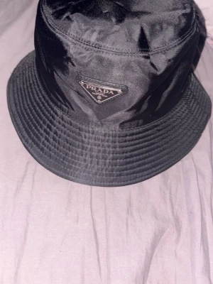 Svart bucket hat från Prada - Snygg svart bucket hat från Prada i glansigt nylonmaterial. Klassisk form med bred brätte och den ikoniska metalltriangeln med Prada-logga framtill. Perfekt accessoar för en trendig streetwear-look.