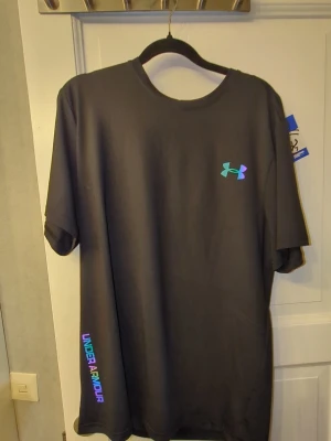 Svart Under Armour t-shirt - Snygg svart t-shirt från Under Armour med regnbågsfärgad logga på bröstet och text längs sidan. Tillverkad i ett mjukt och lätt material som passar perfekt till träning eller chill. Klassisk rund hals och korta ärmar för en clean look.