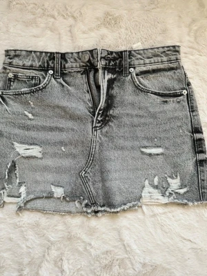Grå jeanskjol - Säljer en grå jeanskjol från Zara i kort modell med rå kant och slitna detaljer framtill och baktill. Säljer kjolen då den inte passar mig 